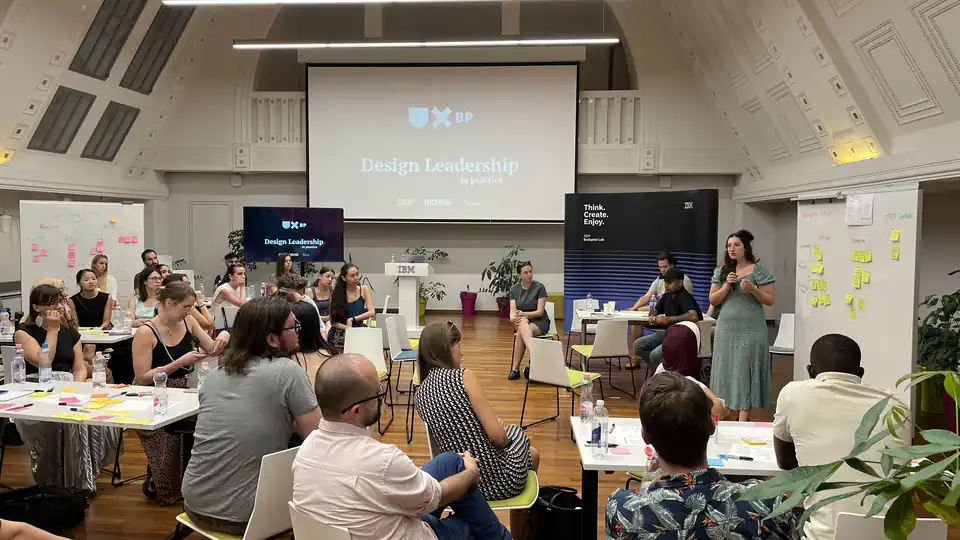 Terem az IBM Budapest Labben: a résztvevők kiscsoportokban ülnek az asztaloknál és cetlikkel dolgoznak, jobb oldalt egy facilitátor beszél. A kivetítőn: Design Leadership in practice.